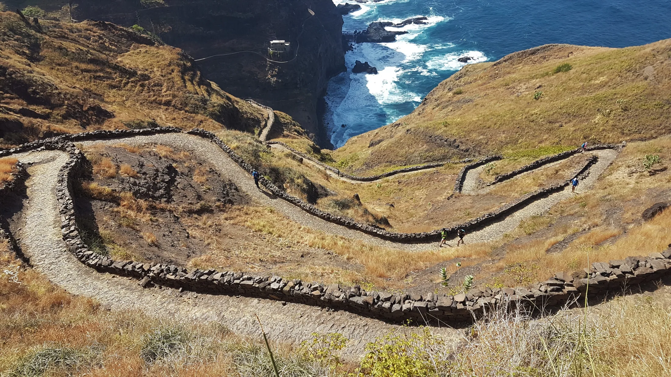 Hikers navigate a steep, winding cobblestone path lined with stone walls that snakes down a dry hillside toward a dramatic coastal cliff where ocean waves crash below.