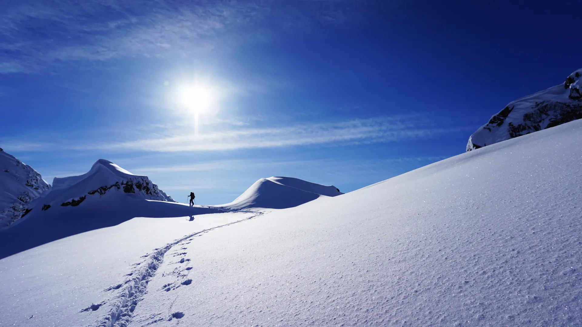 Snowy mountain landscape with a bright sun, a single skier in the distance, and ski tracks winding through the snow.