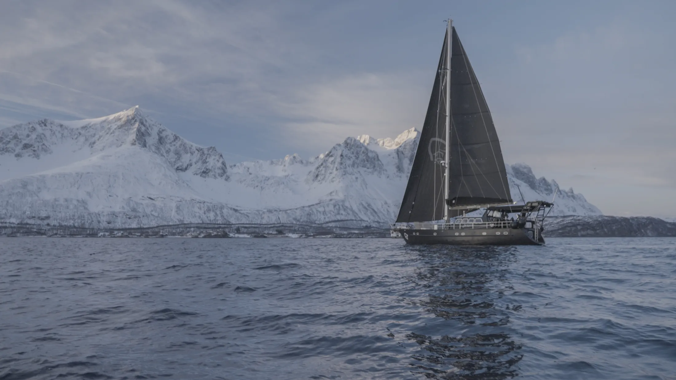 A sailboat with dark sails navigating calm waters with snow-covered mountains in the background.