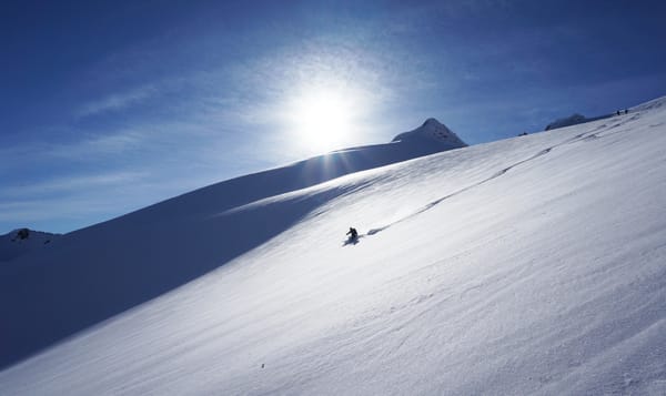 A lone skier on a vast snow-covered mountain slope under a bright sun and blue sky.