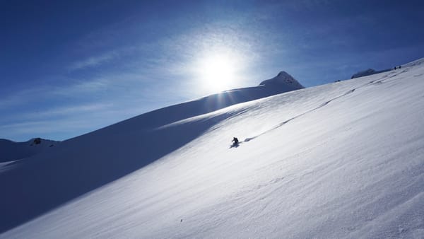 A lone skier on a vast snow-covered mountain slope under a bright sun and blue sky.