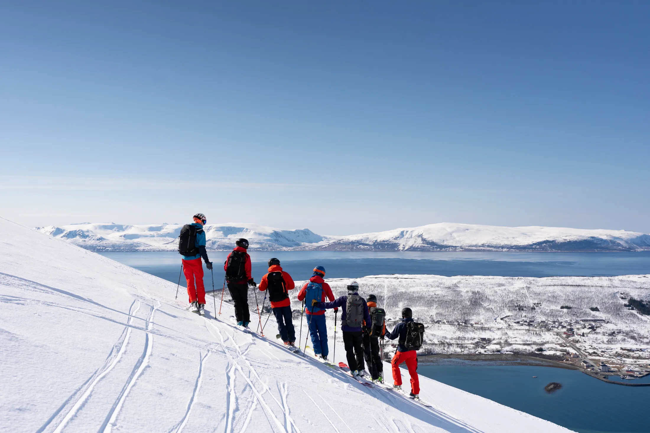 A group of skiers in winter clothing standing on a snow-covered mountain slope overlooking a fjord and ocean under a clear blue sky.