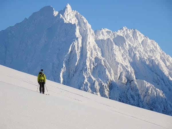 A lone skier in a bright yellow jacket ascends a vast snowy slope with towering, snow-covered peaks under a clear blue sky in the background.