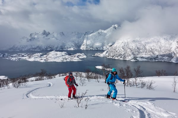 Two ski tourers ascending a snowy slope overlooking a fjord and mountains in Lofoten, Norway.