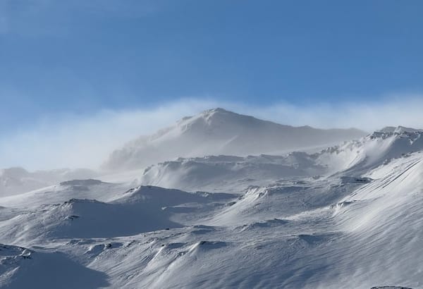 A snowy mountain peak against a blue sky, with wind blowing snow across the summit.