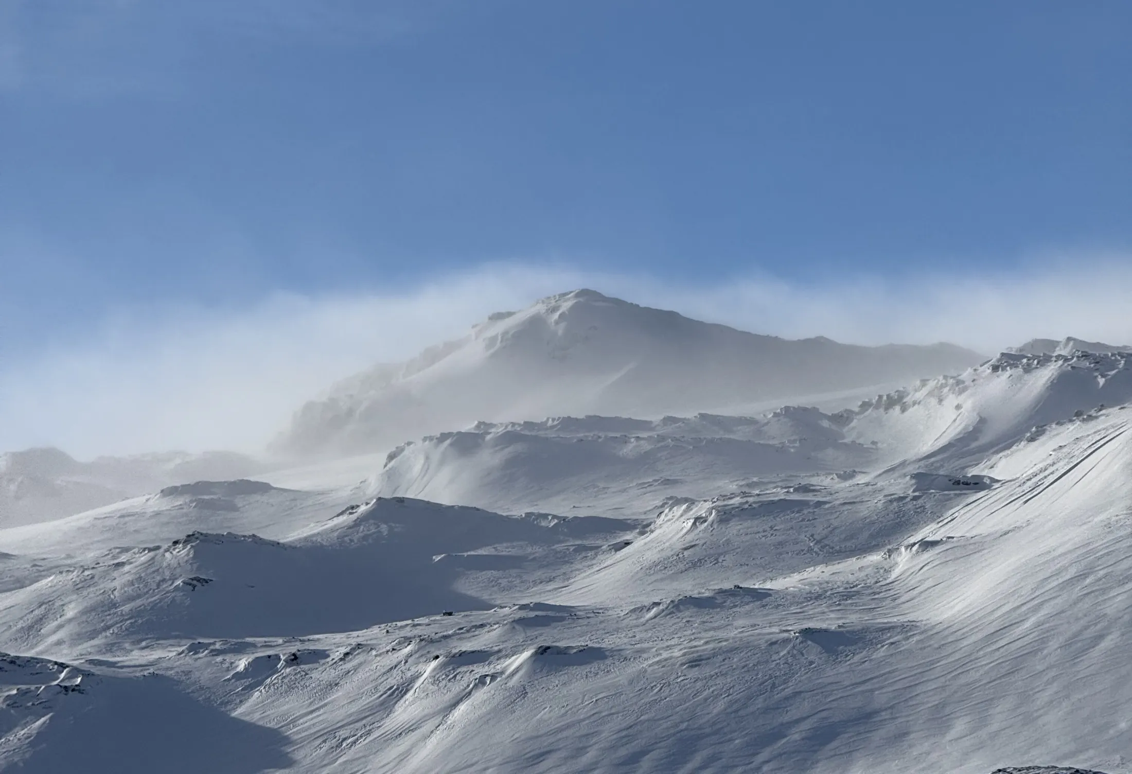 A snowy mountain peak against a blue sky, with wind blowing snow across the summit.