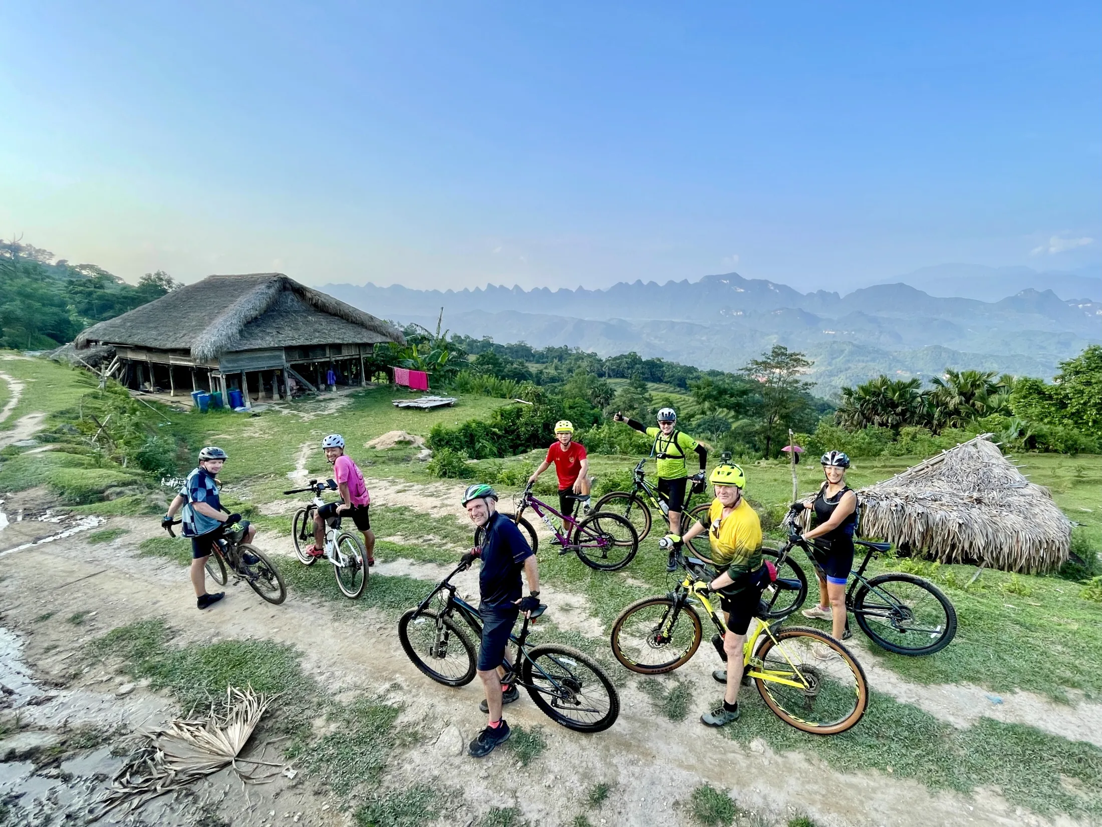 A group of mountain bikers on a dirt path with a scenic mountain view and a traditional house in the background.