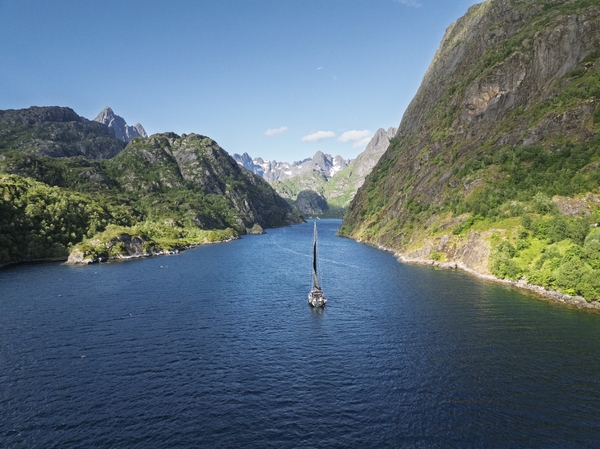 A sailboat navigating a narrow, blue fjord surrounded by steep, green mountains under a blue sky.
