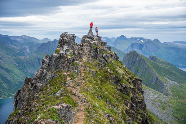 Two hikers standing on a rocky mountain summit overlooking a valley under a cloudy sky.