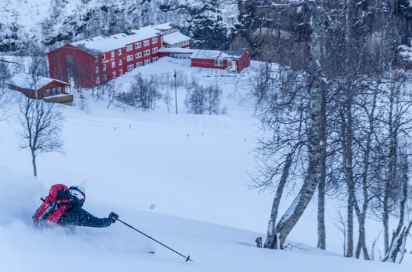 A skier with a red backpack carves through deep powder snow on a slope overlooking a red mountain hotel down in the valley.