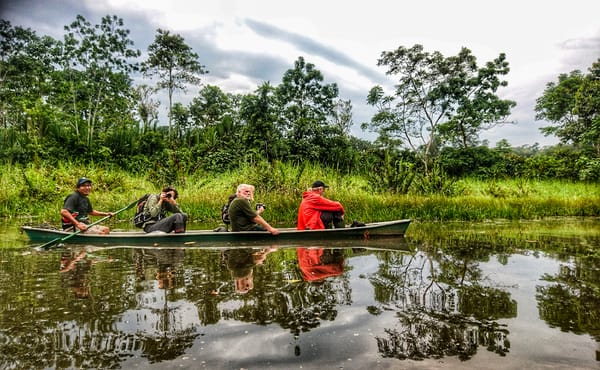 A group of tourists in a canoe rides down a calm river, their reflection mirrored in the water against a background of lush jungle trees under a cloudy sky.