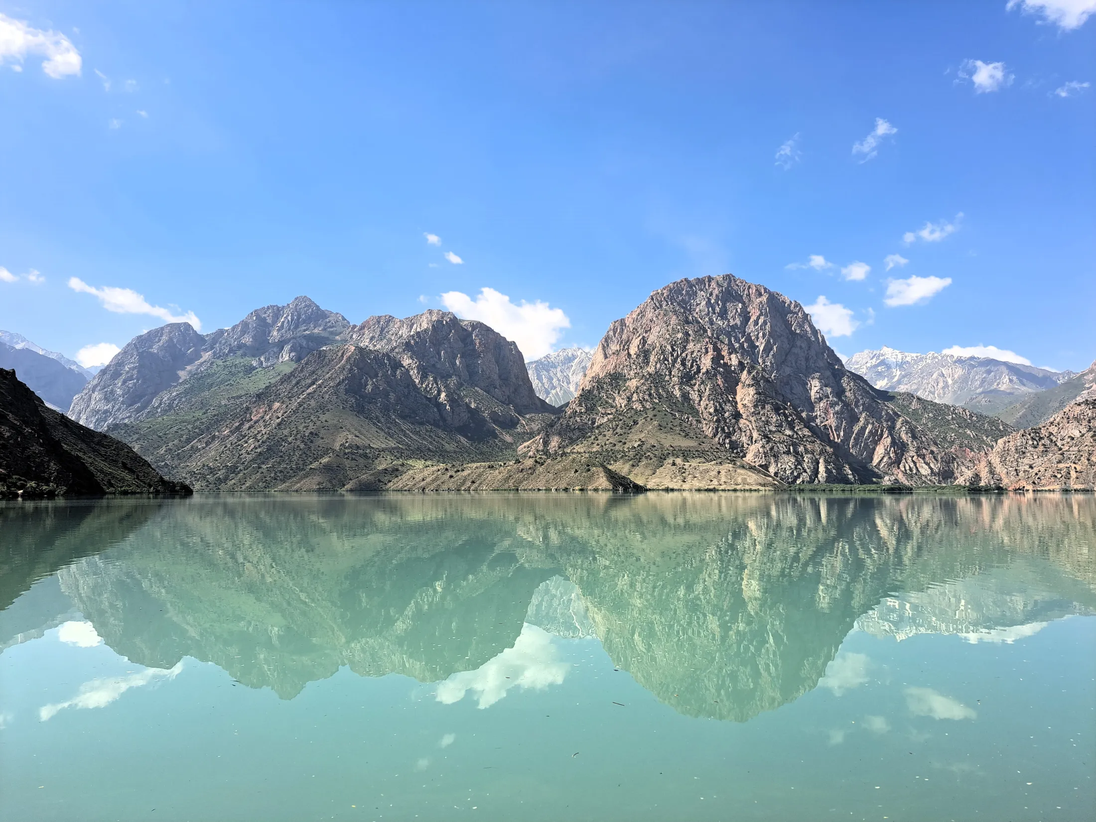 A panoramic view of massive, rocky mountains perfectly mirrored on the glassy surface of a calm, pale turquoise lake under a bright blue sky.
