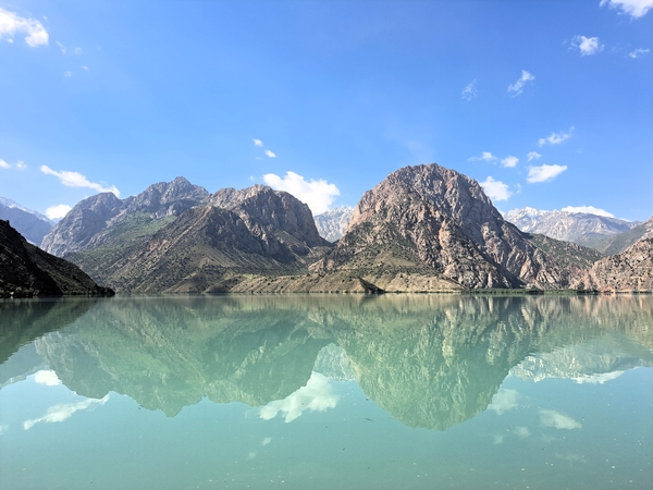 A clear turquoise mountain lake forms a crisp, mirrored reflection of the rugged, barren peaks and blue sky above it.