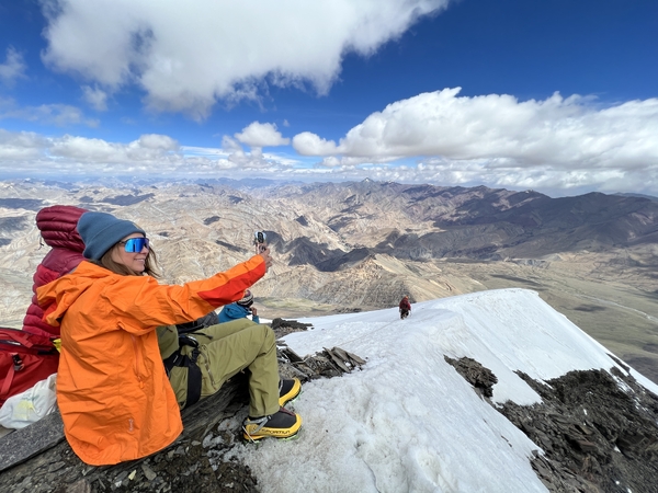 A mountaineer in a bright orange jacket sits on a snowy ridge and takes a selfie, with a vast, rugged mountain landscape stretching out beneath a partly cloudy blue sky.