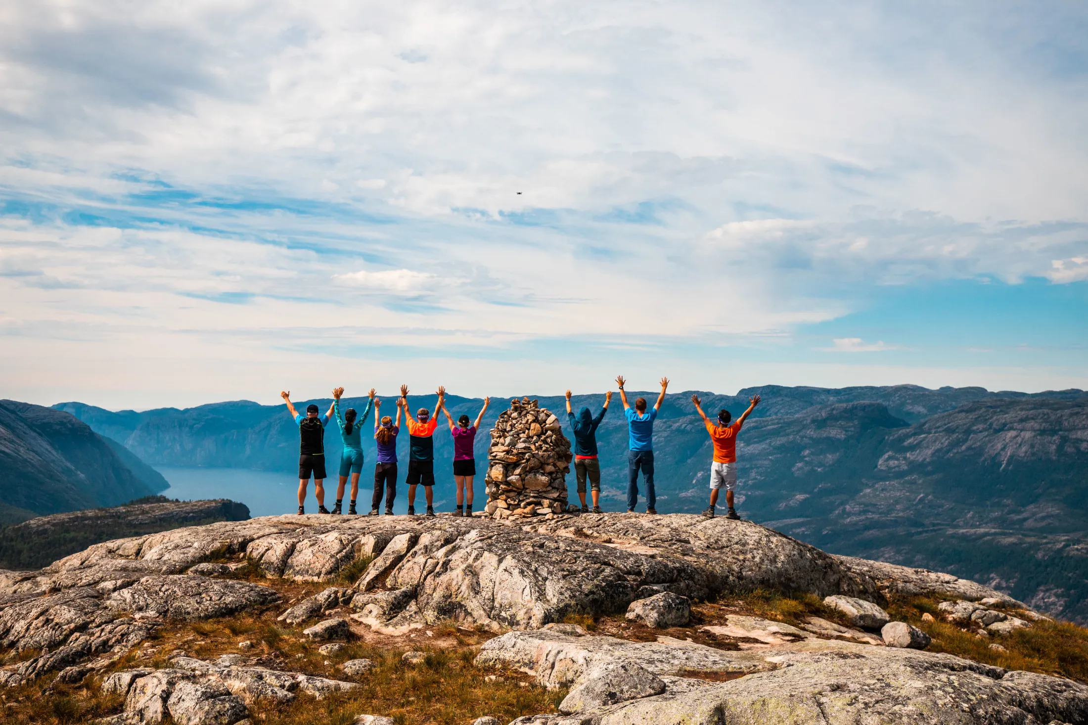 A group on the mountain summit celebrating with raised hands above the fjord.