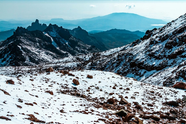 A snowy and rocky alpine slope on Mount Kenya overlooks a rugged valley with dark jagged mountain peaks and blue, hazy ridges stretching into the distance.