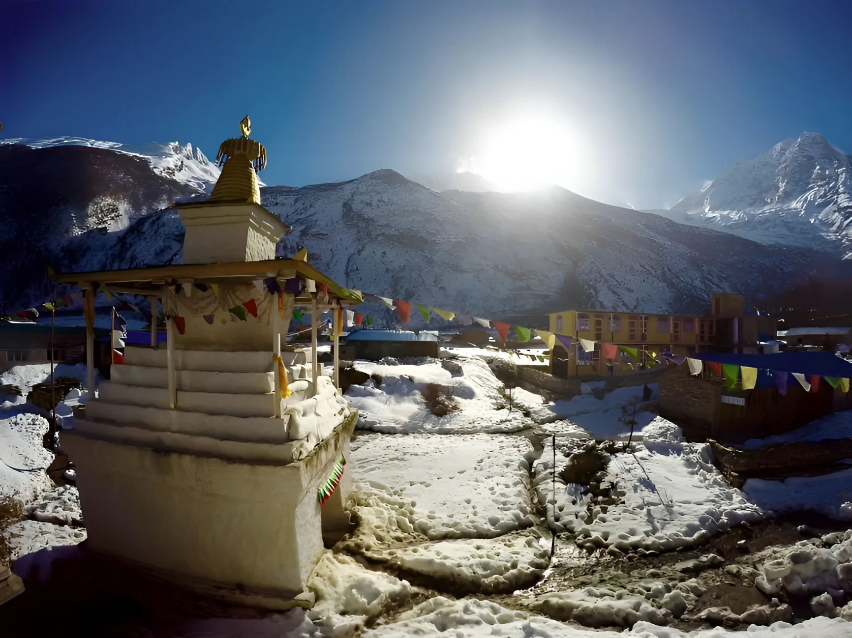 English: A white Buddhist stupa stands in a snow-covered mountain village with colorful prayer flags stretching toward buildings under a bright, glaring sun.