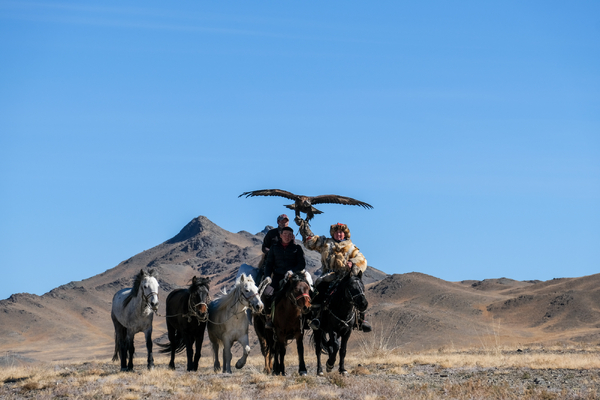 Three individuals ride horses across an arid landscape, with one person in traditional attire holding a large golden eagle aloft with wings spread, set against a backdrop of rolling brown mountains and a clear blue sky.