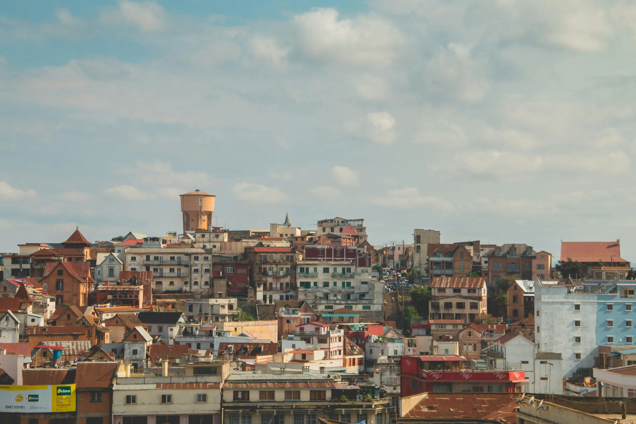 A dense hillside city landscape featuring multi-story buildings and a prominent water tower under a partly cloudy sky.