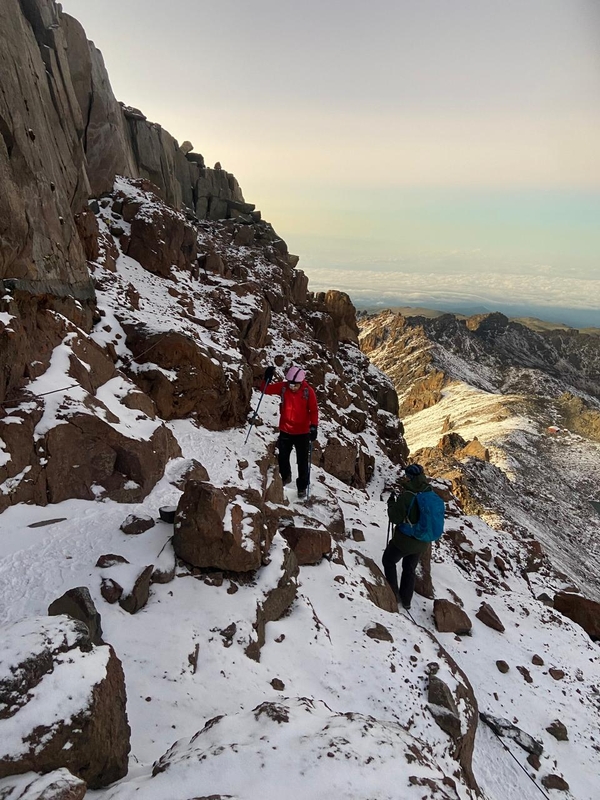 Two hikers navigate a steep, rocky, and snow-covered mountain ridge on Mount Kenya under a pale sky.