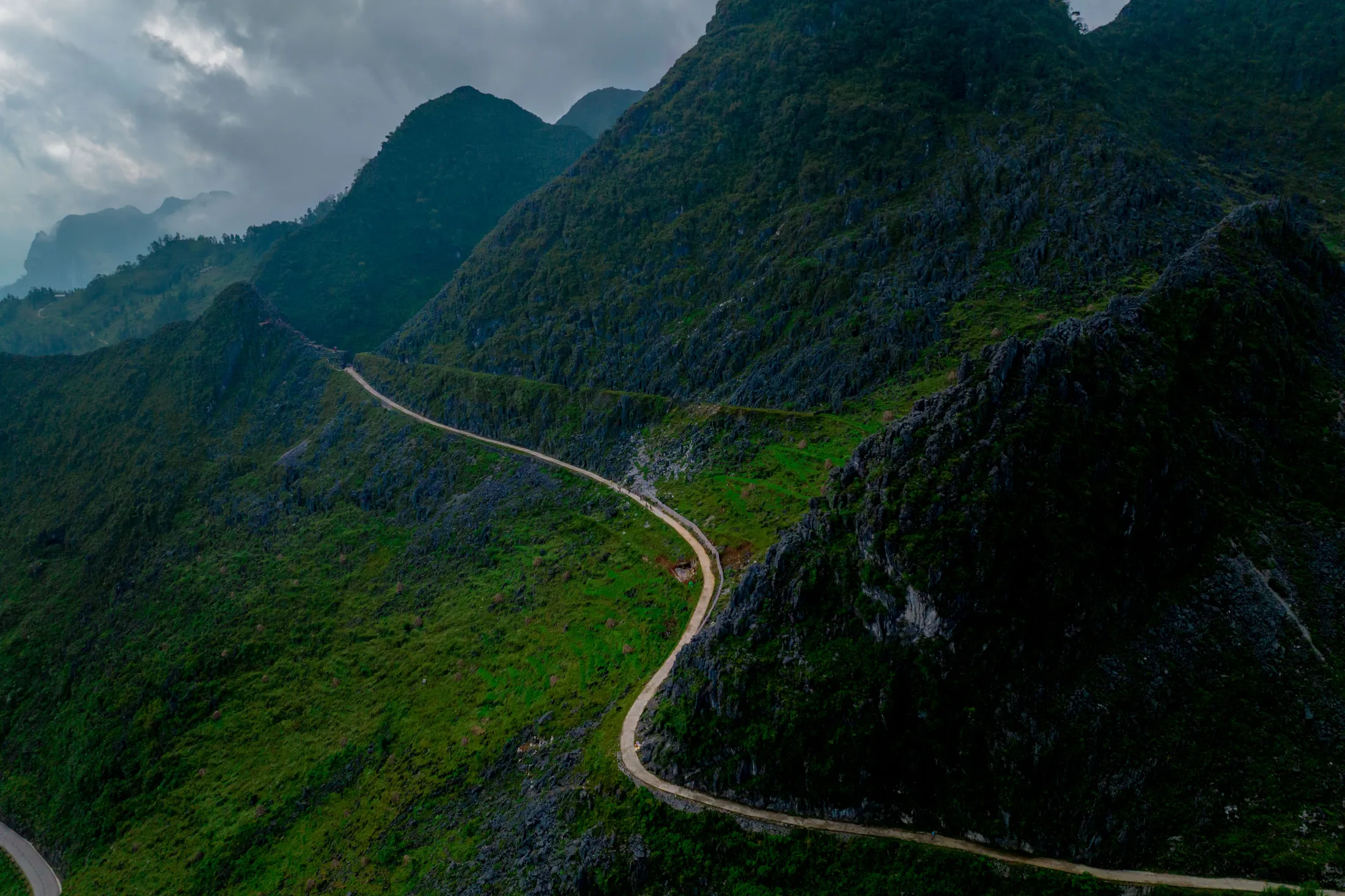An aerial view of a winding road cutting through steep, green mountain slopes under a cloudy sky.