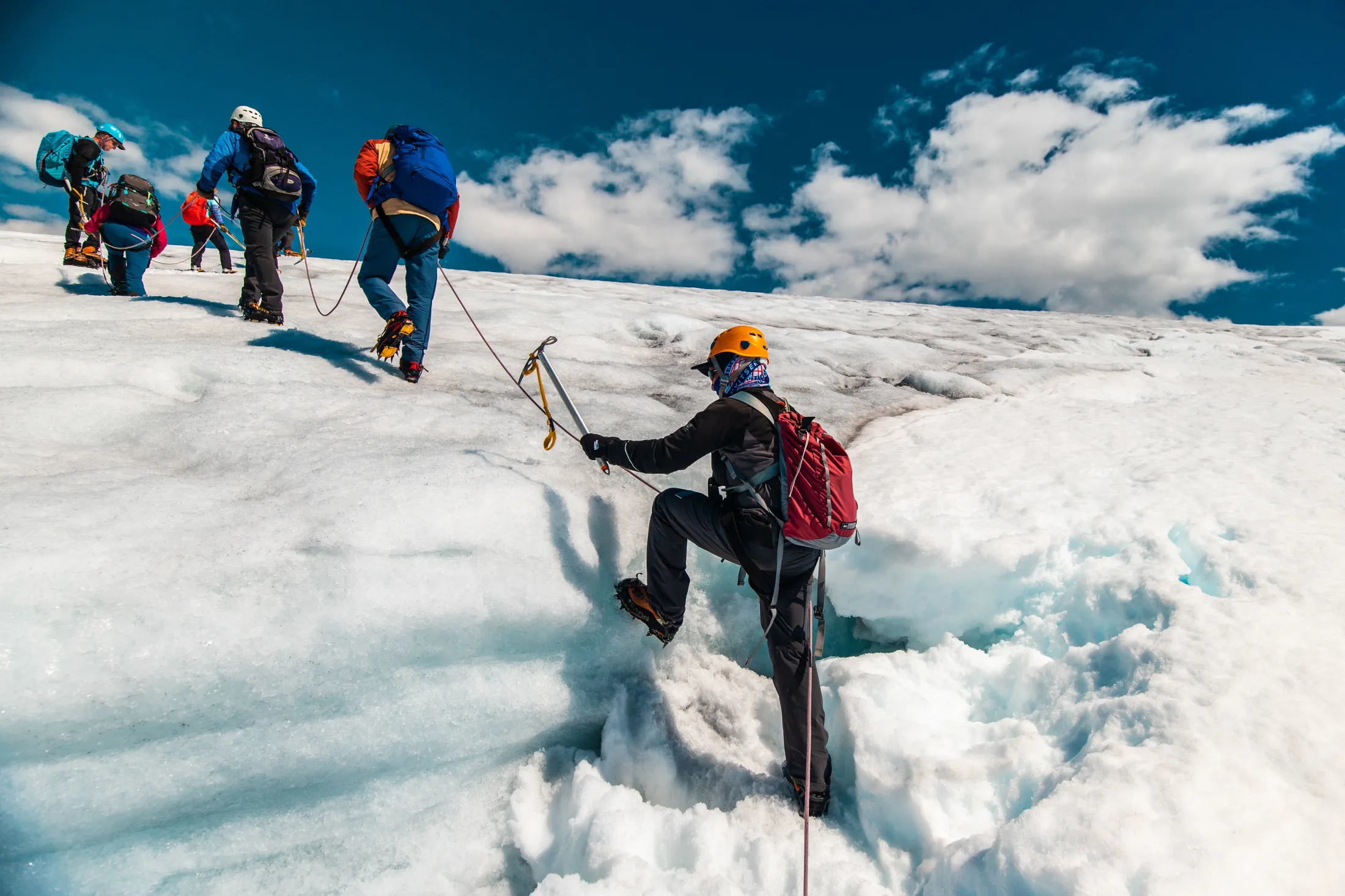 A group of roped mountaineers climbing a glacier under a partly cloudy blue sky.