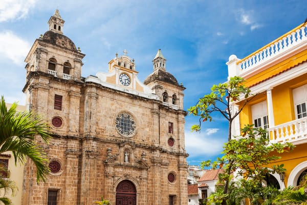 Facade of the historic Church of San Pedro Claver in Cartagena, Colombia, with a bright yellow building on the right under a blue sky.