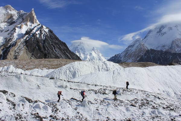 A small group of trekkers hikes across a vast, icy glacier landscape framed by towering snow-covered peaks under a bright blue sky.