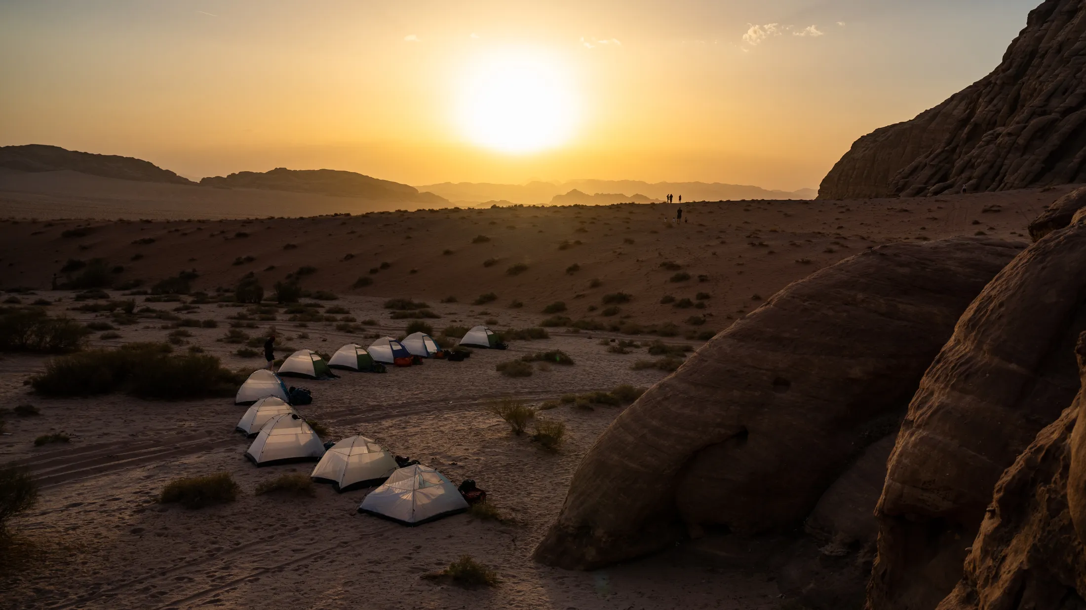 A row of tents pitched on golden sand in a desert landscape during a glowing sunset.