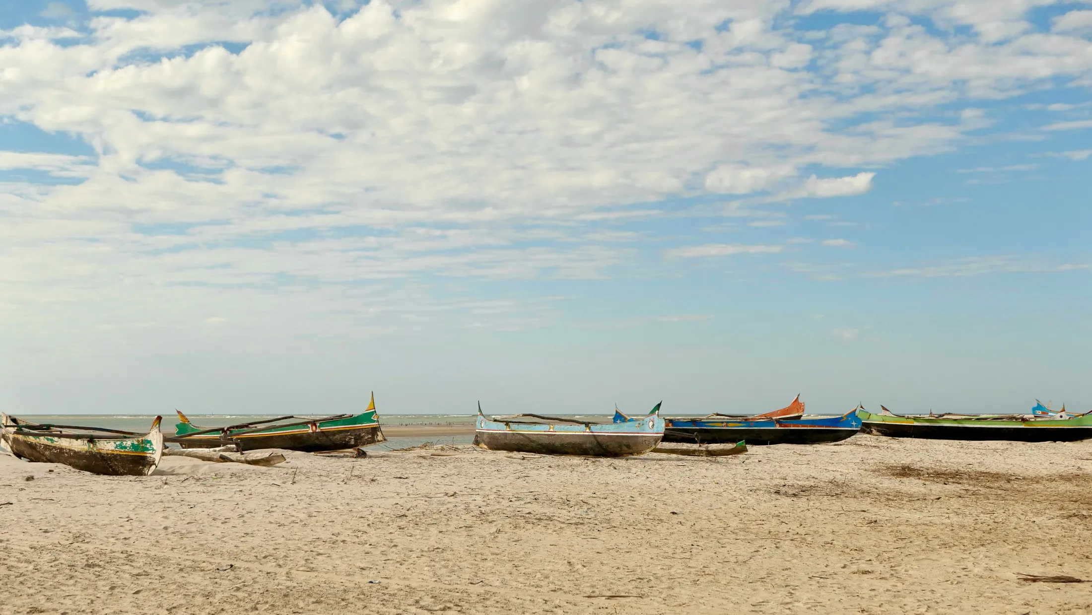 Several colorful wooden pirogues rest on a sandy beach looking out toward the ocean under a cloud-filled sky.
