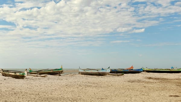 Several colorful wooden pirogues rest on a sandy beach looking out toward the ocean under a cloud-filled sky.
