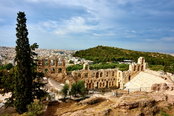An elevated view captures the ancient stone ruins of the Odeon of Herodes Atticus amphitheater in Athens, with a lush green hill and a sprawling cityscape stretching out beneath a cloudy blue sky.