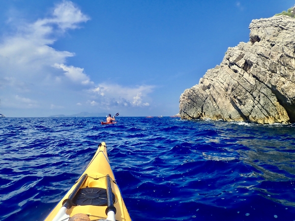 A first-person view from a yellow kayak paddling on choppy, deep blue sea water, following another kayaker toward a rugged, rocky cliffside under a bright blue sky with scattered clouds.