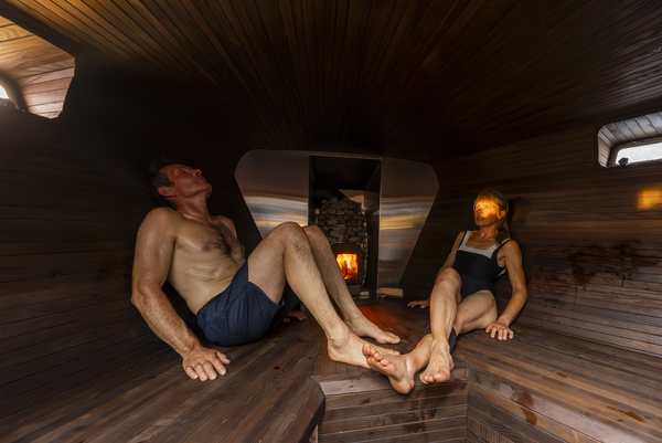 A man and a woman relaxing in a rustic, dark-wood sauna with a view of the outside.
