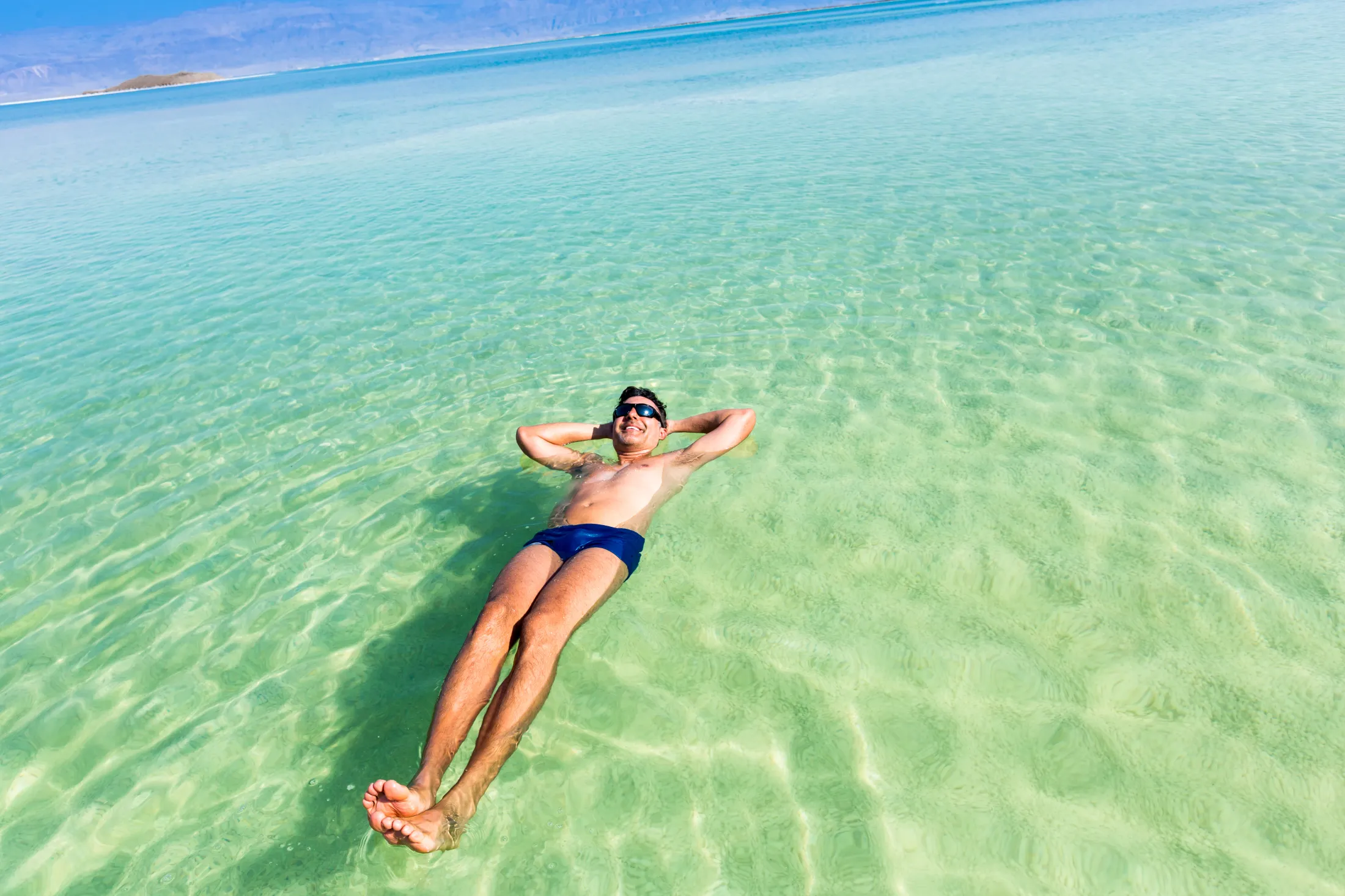 A man floating relaxed on his back in the crystal-clear, turquoise water of the Dead Sea.