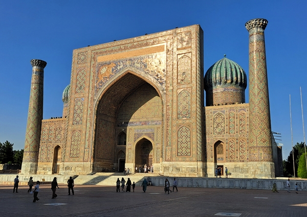People walk in front of a massive, ancient madrasah building adorned with intricate tile work, a large central archway, prominent domes, and tall minarets under a clear blue sky.