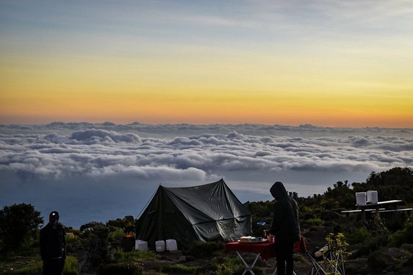 A mountain campsite sits high above a sea of thick clouds as the sun sets with a warm orange gradient in the sky.