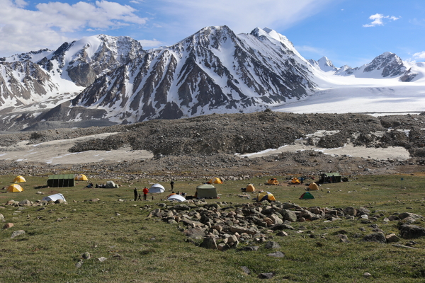 A campsite with several colorful tents and scattered people is set up on a grassy, rocky field at the base of a massive, snow-covered mountain range under a blue sky.