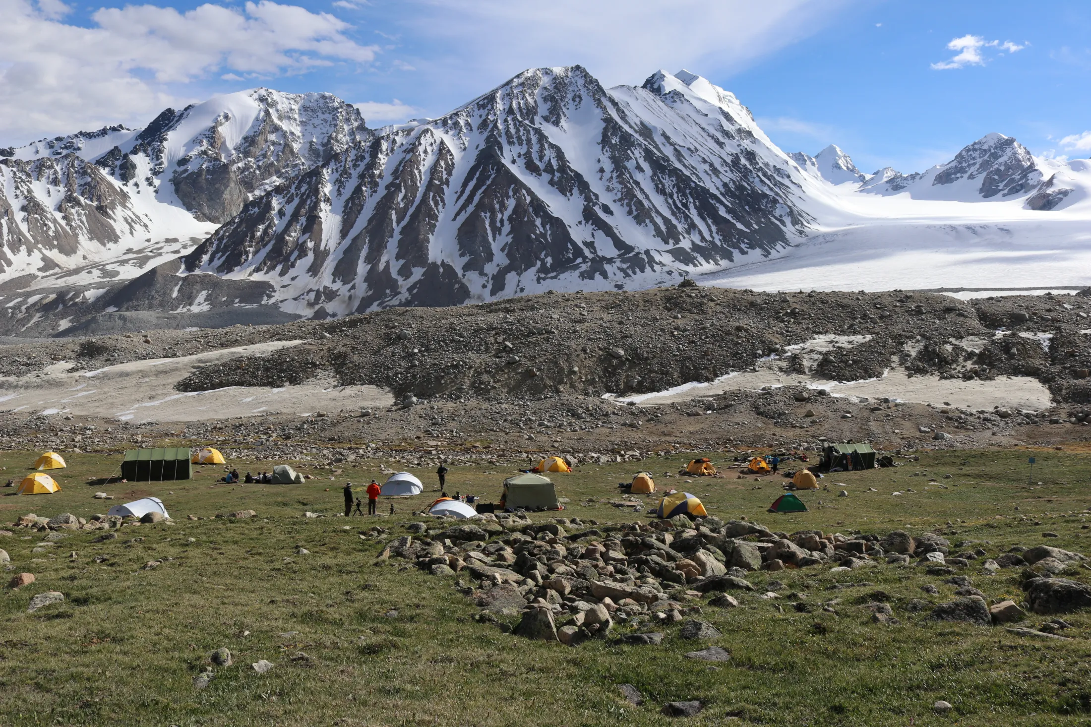A collection of brightly colored tents and a few hikers are scattered across a green, rocky meadow below a massive snow-capped mountain range and an icy glacier under a blue sky.