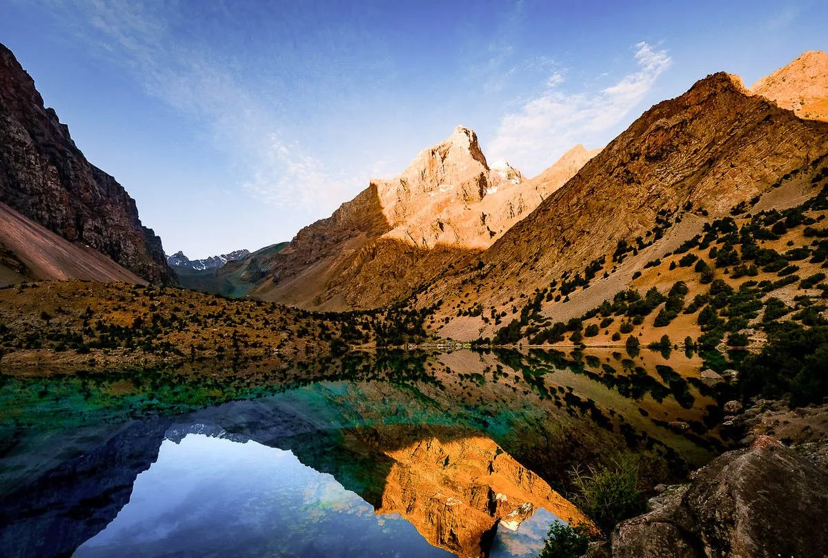 A still, clear alpine lake creates a perfect reflection of a towering mountain peak illuminated by sunlight and surrounded by rugged, tree-dotted slopes.