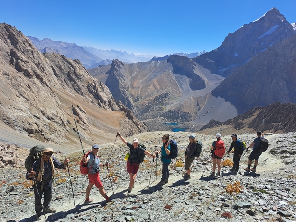 A group of hikers poses along a rocky mountain trail overlooking a deep valley and a vast landscape of towering, rugged peaks under a clear blue sky.