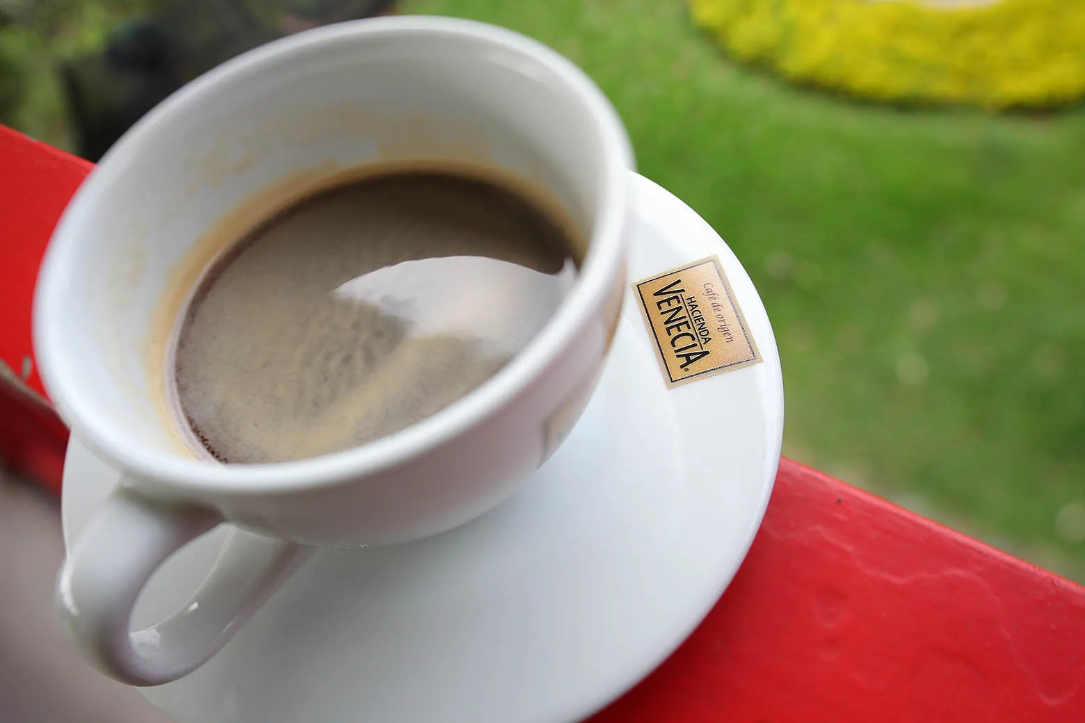 A white cup of black coffee on a matching saucer, resting on a red railing with a blurred green background.