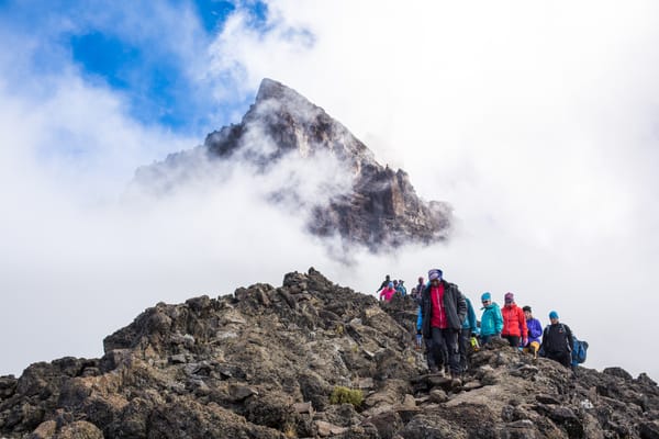 A line of hikers walks down a rocky mountain ridge as low-hanging clouds surround a sharp peak rising in the background.