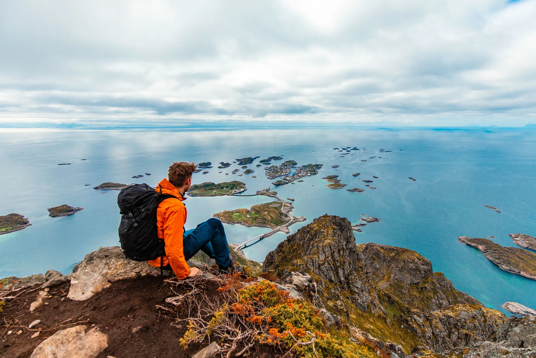 A hiker wearing an orange jacket and a black backpack sits on a mountaintop, looking out at islands scattered across a vast ocean under a cloudy sky.