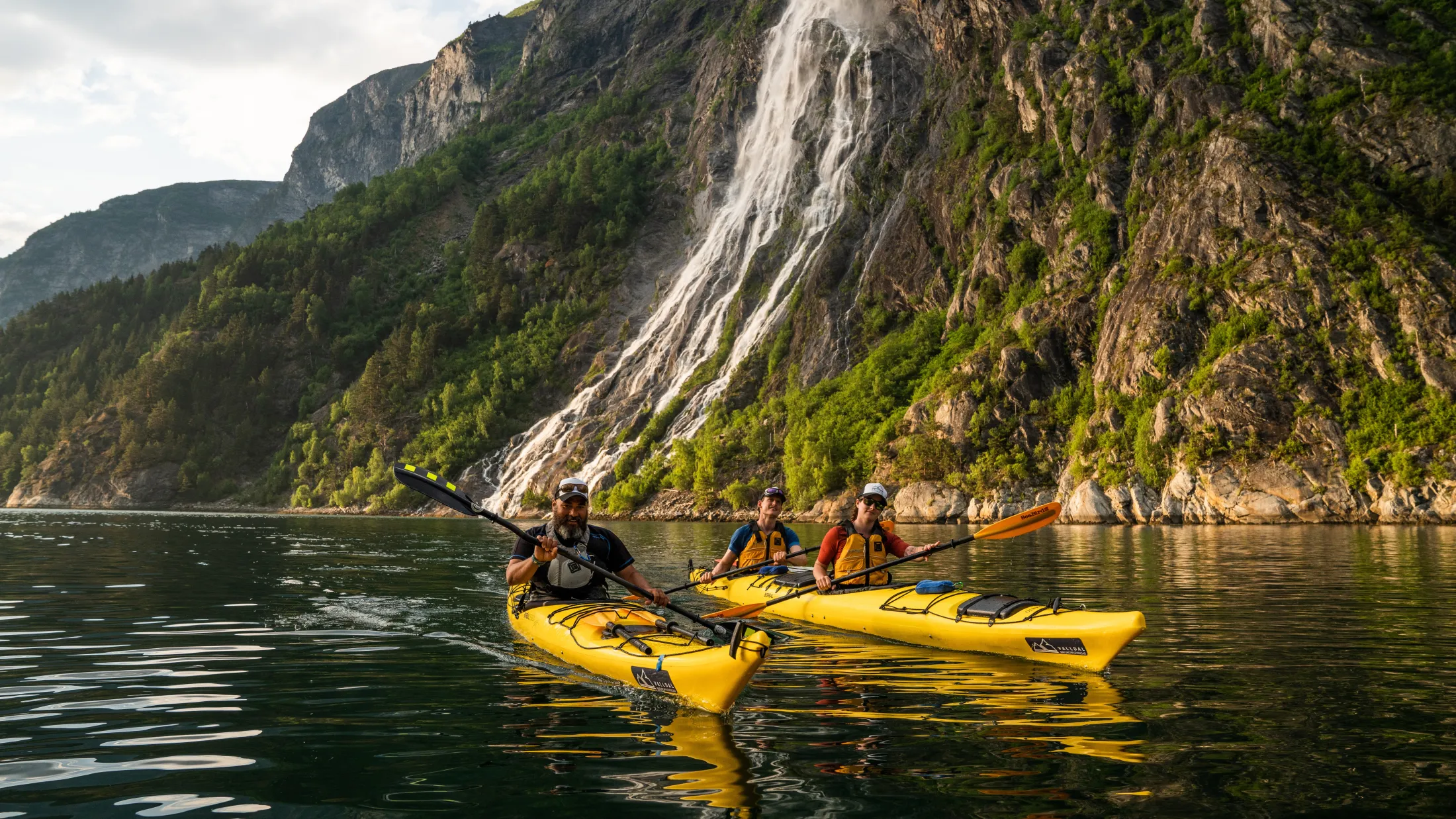 Two people kayaking in a Norwegian fjord with a large waterfall and steep cliffs in the background.