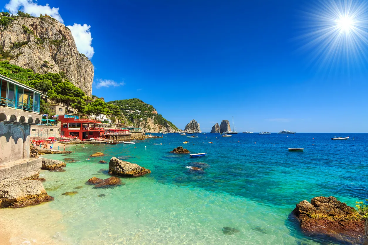 A sunny coastal landscape featuring turquoise water, rocky cliffs, and buildings along the shore, with the bright sun shining in a clear blue sky.