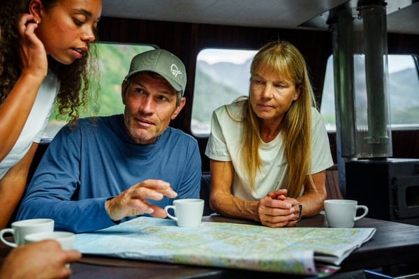 A man and two women look at a map together inside a boat cabin, with white cups on the table.