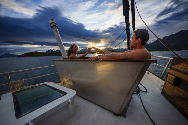 A man and woman relaxing in a hot tub on the deck of a boat, with a dramatic cloudy sky and sea in the background.