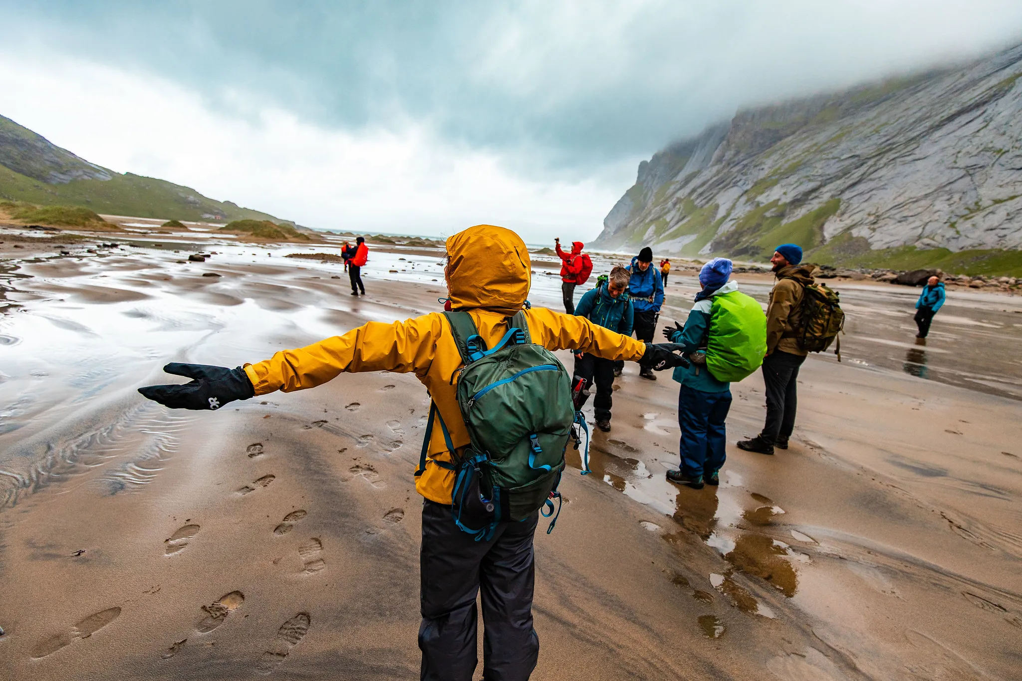 A hiker with arms outstretched in a yellow jacket, with other hikers on a muddy beach under a cloudy sky.