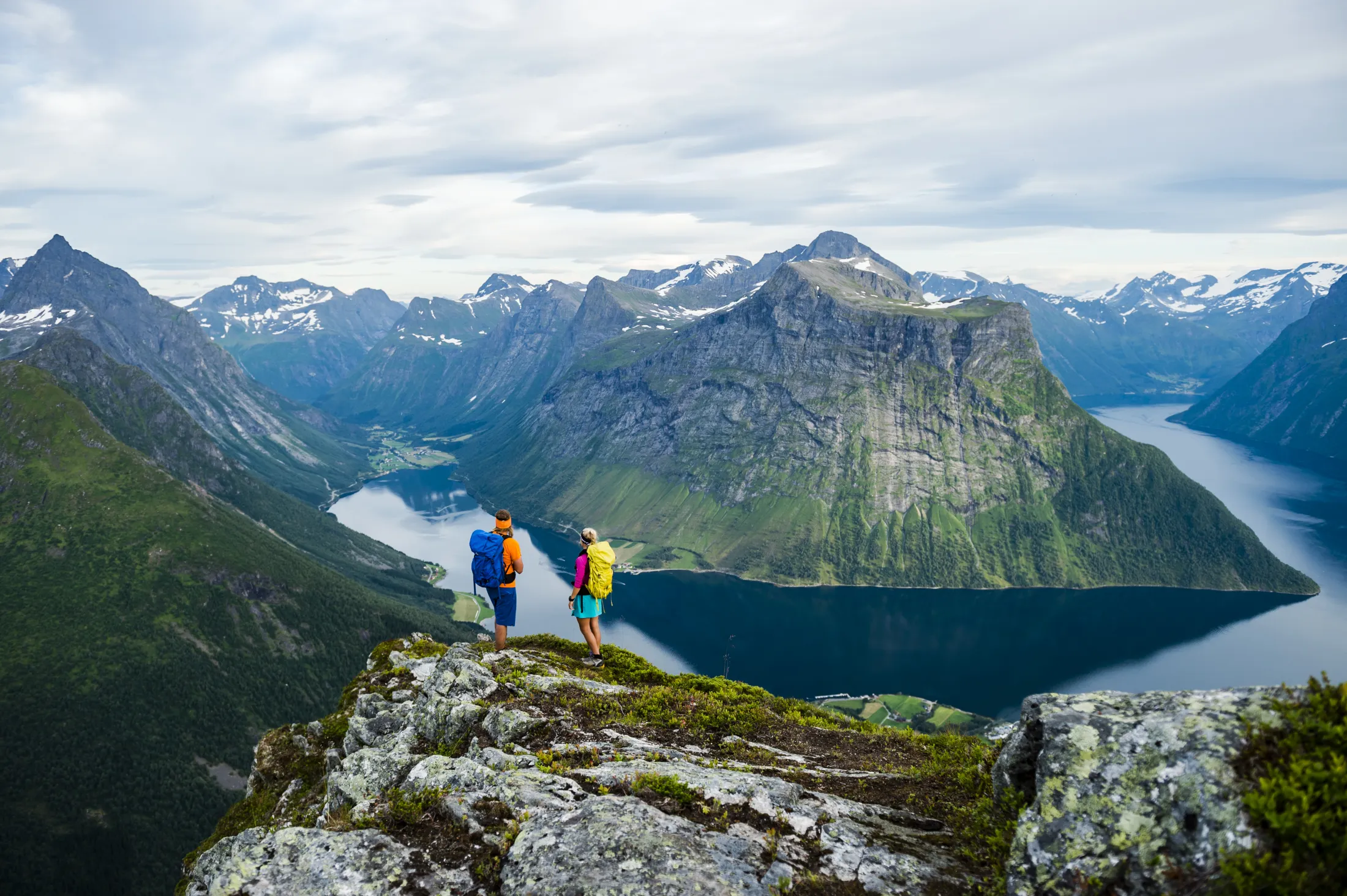 Two hikers standing on a mountain ridge looking down at a fjord surrounded by steep peaks.
